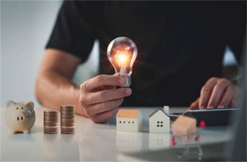 person holding a dimly lit light globe in the background, with a small piggy bank and stacks of coins on a table in front of them