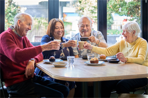 Group of four older people seated in a cafe, cheering with cups of coffee